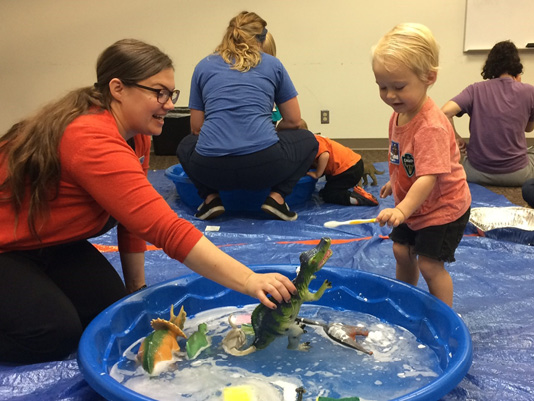 A librarian and a child play with dinosaur dolls in a kiddie pool.